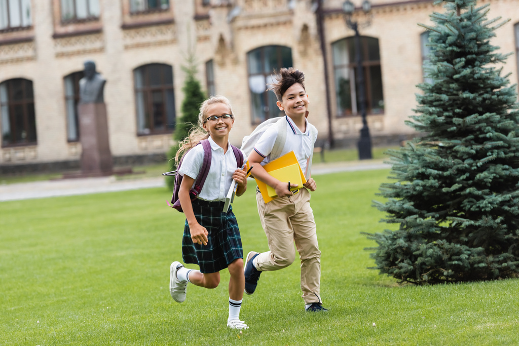 Uśmiechnięte wielonarodowe dzieciaki z notatnikami biegającymi po trawniku w parku  - Zdjęcie, obraz