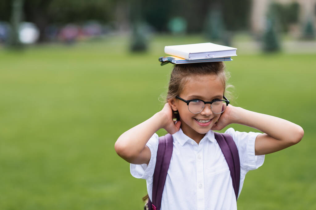 Glimlachend schoolmeisje in bril met notitieboekjes op het hoofd buiten  - Foto, afbeelding