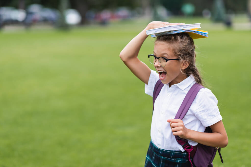Nadšený školák s batohem drží kopie knih na hlavě venku  - Fotografie, Obrázek