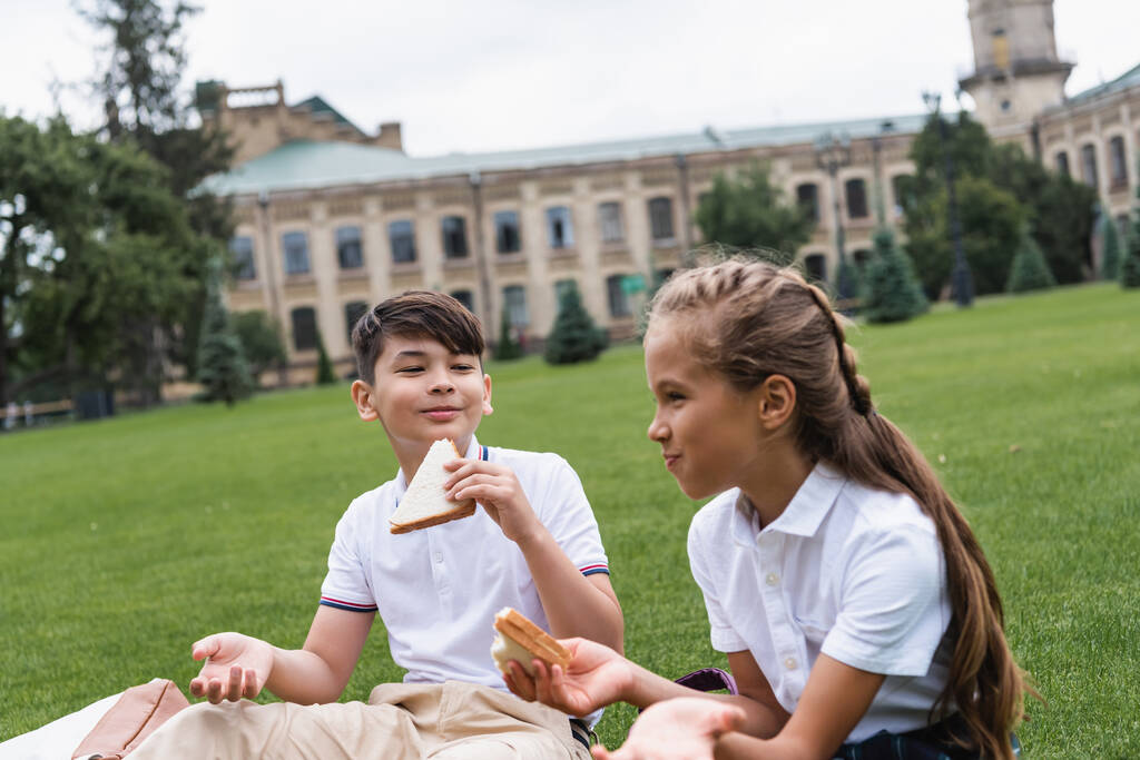 Mosolygó ázsiai iskolás kezében szendvics közel barátja gyep a parkban  - Fotó, kép