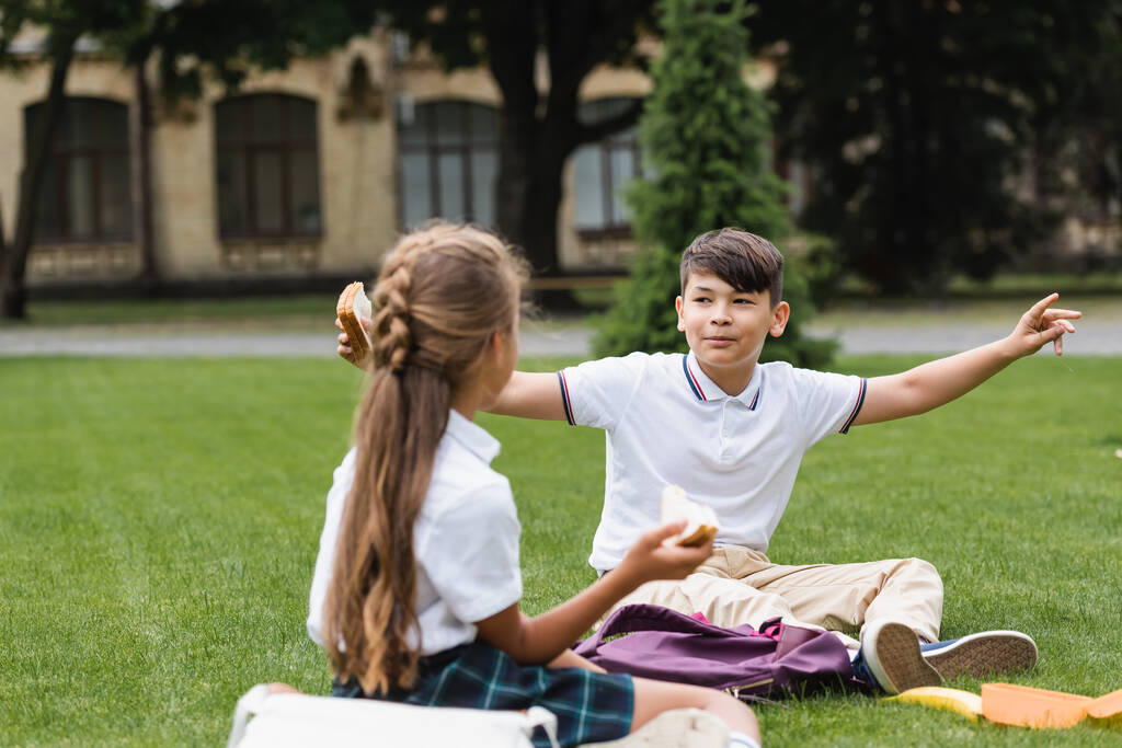 Asiático estudante segurando sanduíche e falando com amigo turvo na grama no parque  - Foto, Imagem