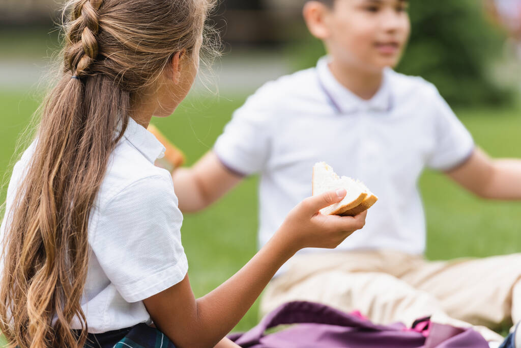 Preteen schoolgirl holding sandwich near blurred asian friend in park  - 写真・画像