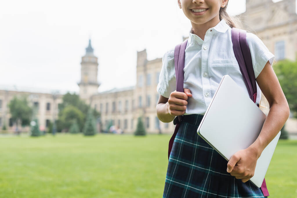 Gesneden weergave van glimlachende tiener schoolmeisje met laptop en rugzak buiten  - Foto, afbeelding