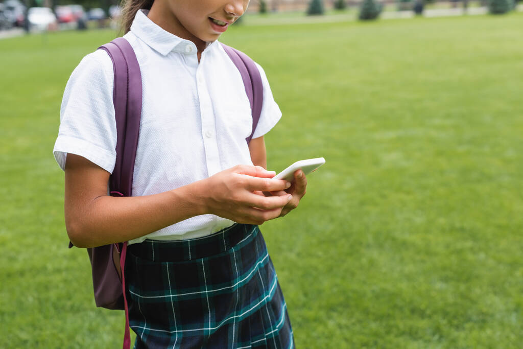 Oříznutý pohled na usmívající se školák s batohem pomocí smartphonu v parku  - Fotografie, Obrázek