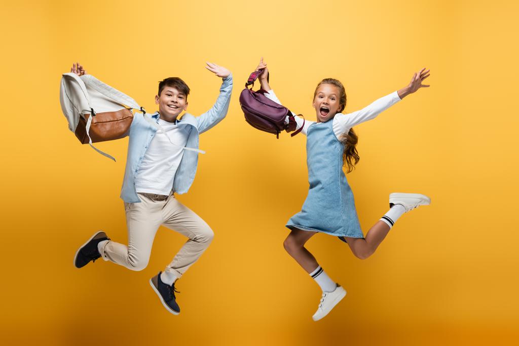 Sırt çantaları sarı arka planda zıplayan heyecanlı çoklu etnik çocuklar. - Fotoğraf, Görsel