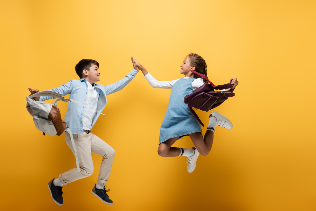 Side view of multiethnic schoolkids with backpacks giving high five while jumping on yellow background - Photo, Image