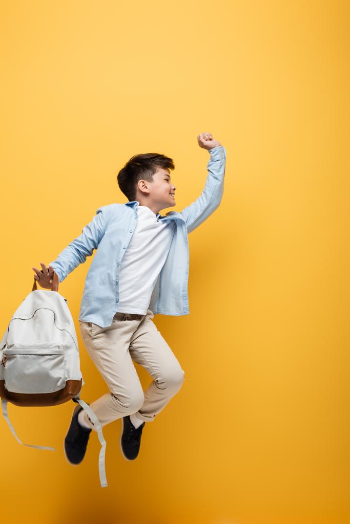 Vue latérale de l'écolier asiatique tenant le sac à dos et sautant isolé sur jaune  - Photo, image