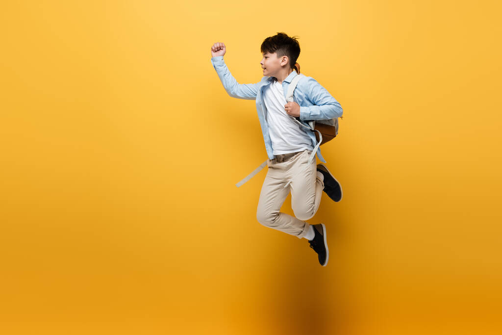 Side view of asian schoolkid with backpack jumping on yellow background - Photo, Image