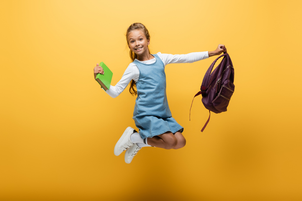Escolar sonriente con mochila y libro saltando sobre fondo amarillo - Foto, Imagen