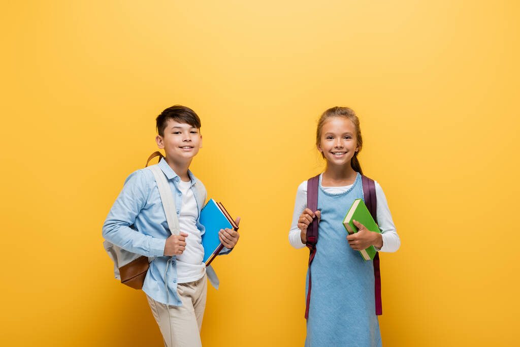 Улыбающиеся межрасовые школьники с книгами и рюкзаками на желтом фоне - Фото, изображение