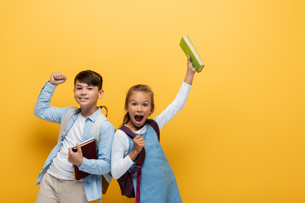 Excited interracial schoolkids holding books and showing yes gesture on yellow background - Photo, Image