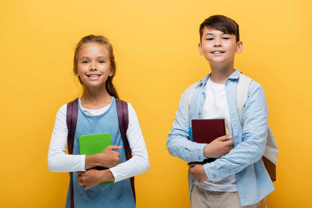 Fröhliche multiethnische Vorschulkinder, die Bücher in der Hand halten und auf gelb isoliert in die Kamera schauen  - Foto, Bild