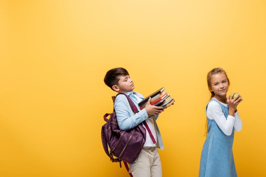 Asian schoolboy with backpack holding books near friend with apple isolated on yellow  - Photo, Image