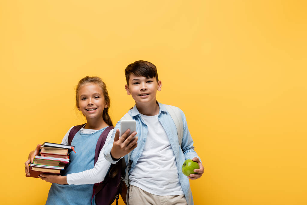 Alegre interracial colegiales sosteniendo libros y teléfono inteligente aislado en amarillo  - Foto, Imagen