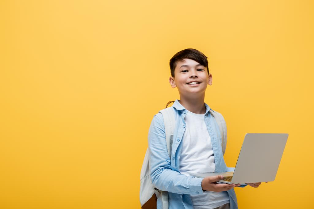 Sırıtkan Asyalı okul çocuğu dizüstü bilgisayarı sarıda yalnız tutuyor.  - Fotoğraf, Görsel