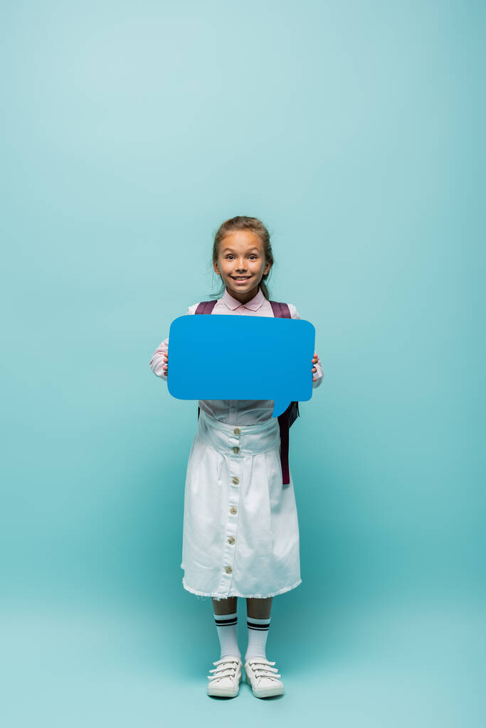 Positive schoolchild with backpack holding speech bubble on blue background  - Photo, Image
