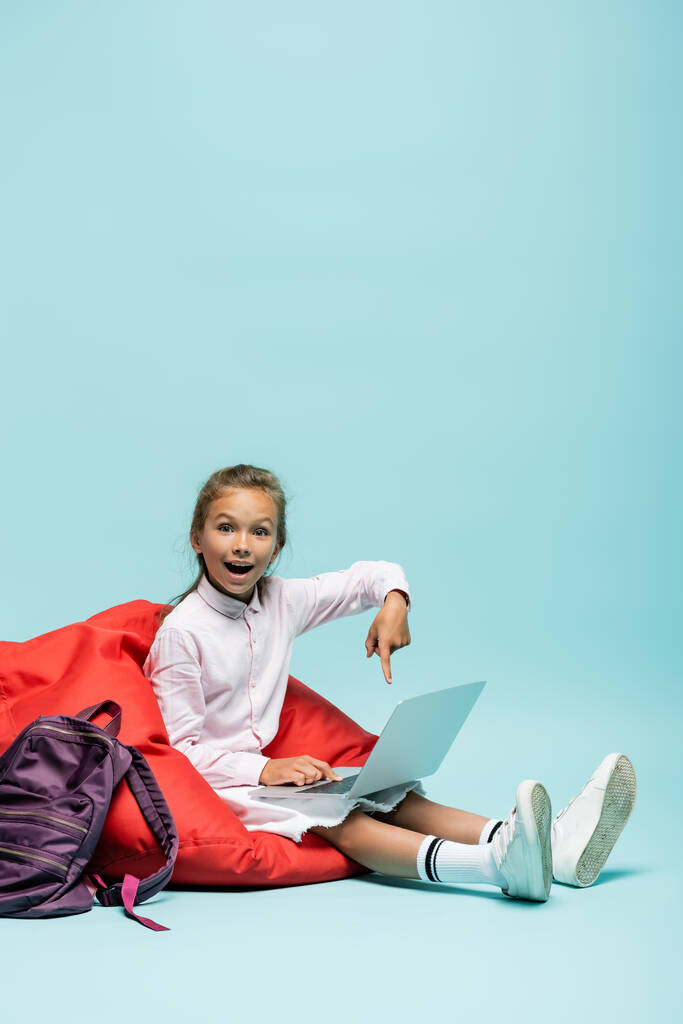 Positive Schulkind zeigt mit dem Finger auf Laptop in der Nähe Rucksack auf blauem Hintergrund  - Foto, Bild