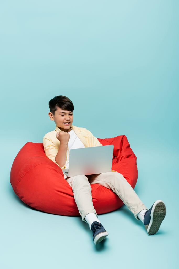 Innoissaan aasialainen koululainen osoittaa kyllä ele, kun käytät kannettavaa tietokonetta papupussi tuoli sinisellä pohjalla  - Valokuva, kuva