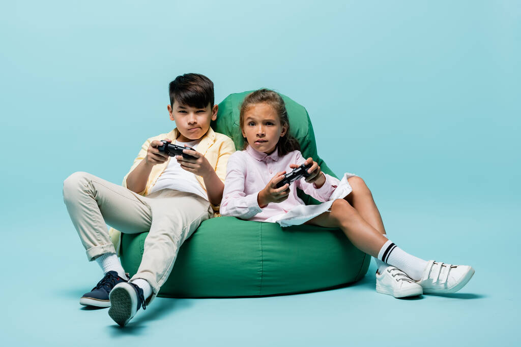 KYIV, UKRAINE - JULY 2, 2021: Focused interracial kids playing video game on beanbag chair on blue background  - Photo, Image