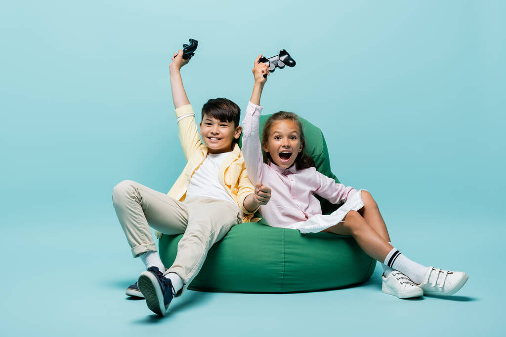 KYIV, UCRANIA - 2 de julio de 2021: Niños interracial excitados sosteniendo joysticks en una silla de frijoles sobre fondo azul  - Foto, imagen