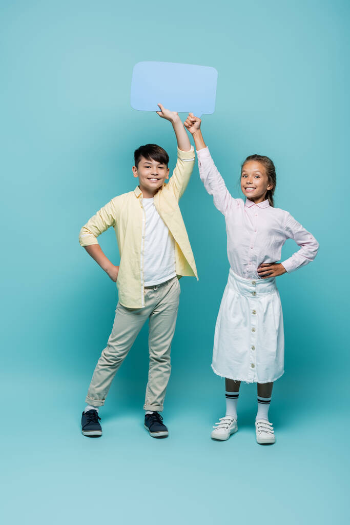 Smiling multiethnic schoolkids holding speech bubble on blue background  - Photo, Image
