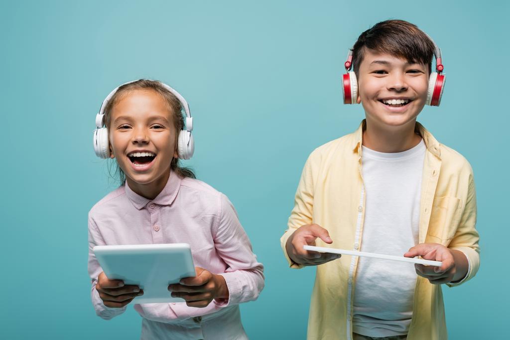 Joyeuses élèves interracial dans des écouteurs tenant des tablettes numériques isolées sur bleu  - Photo, image
