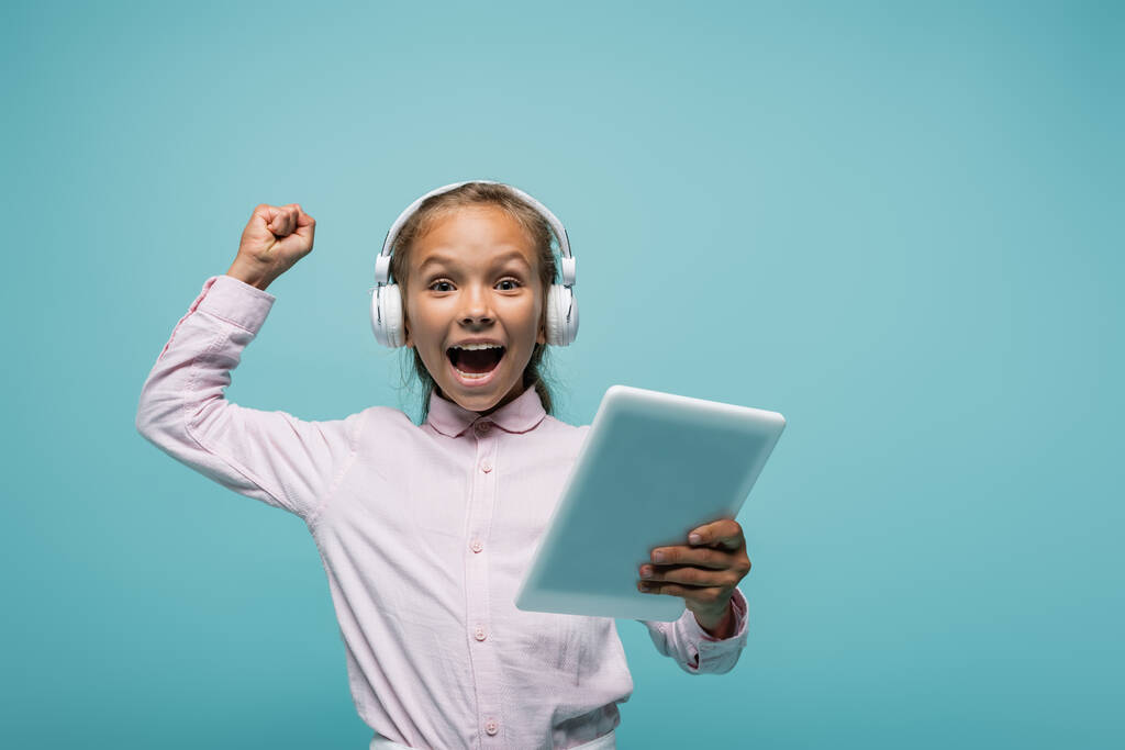 Izgalmas iskolás lány a fülhallgatóban digitális tabletta izolált kék  - Fotó, kép