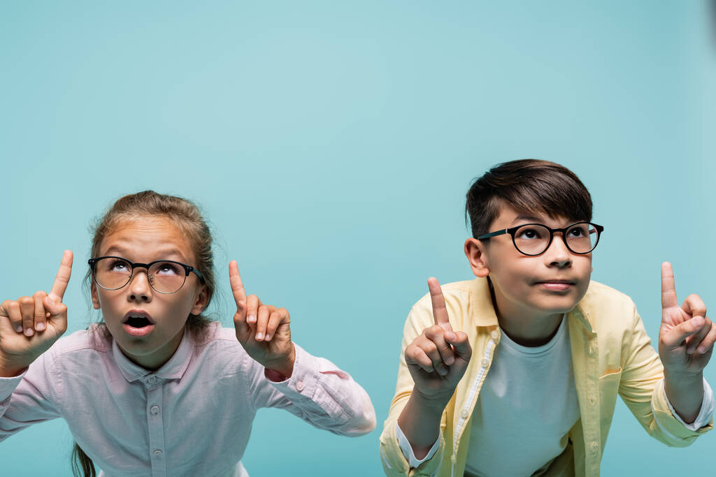 Interracial schoolkids in eyeglasses pointing with fingers isolated on blue  - Photo, Image