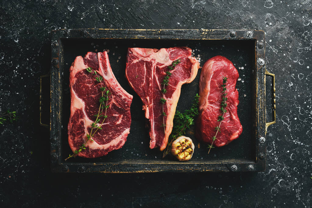 Różnorodność surowych steków wołowych do grillowania z przyprawami: T-bone, striploin steak, ribeye. Widok góry. Na czarnym kamiennym tle. - Zdjęcie, obraz