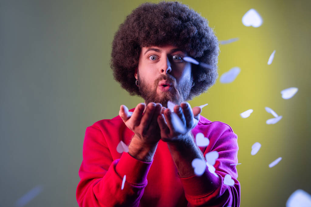 Muotokuva hipster mies Afro kampauksen puhaltaa sydämen muotoinen konfetti, nauttia syntymäpäivä tai Ystävänpäivä, yllään punainen huppari. Sisäuima studio ampui eristetty värikäs neon valo tausta. - Valokuva, kuva