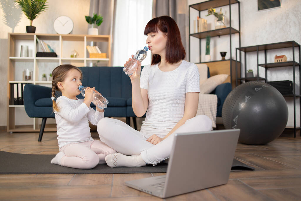 Fiatal anya és lánya sportolnak laptoppal a padlón otthon, miközben friss vizet isznak. Sportos anya gyermekes lánnyal edz reggel. Egészséges családi életmód. - Fotó, kép