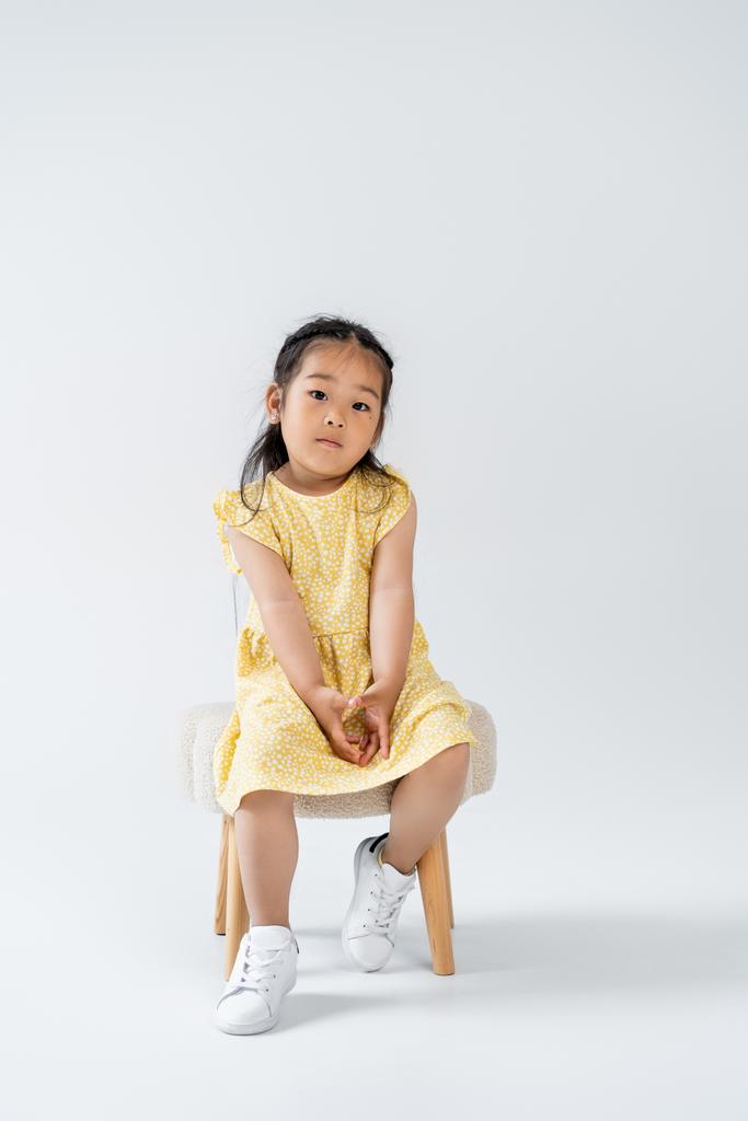 tutta la lunghezza del bambino asiatico in abito giallo e scarpe da ginnastica bianche seduto sulla sedia su grigio - Foto, immagini
