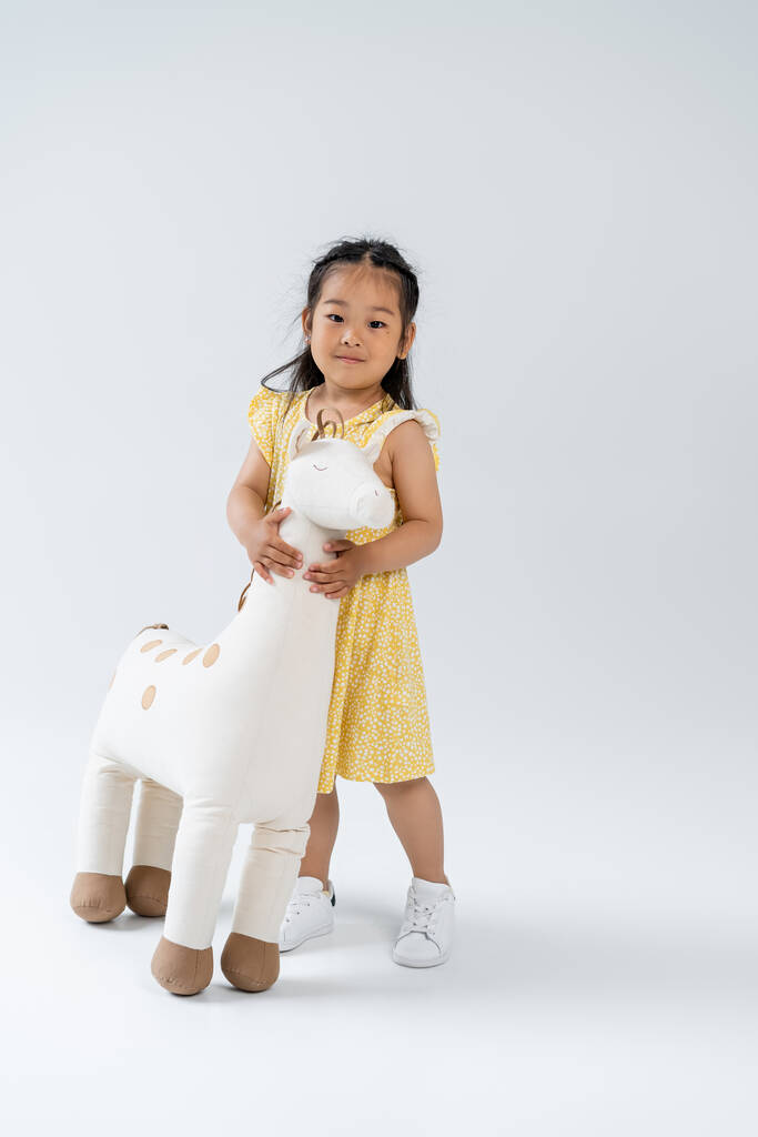 volledige lengte van blij aziatisch kind houden speelgoed giraffe en staan op grijs - Foto, afbeelding