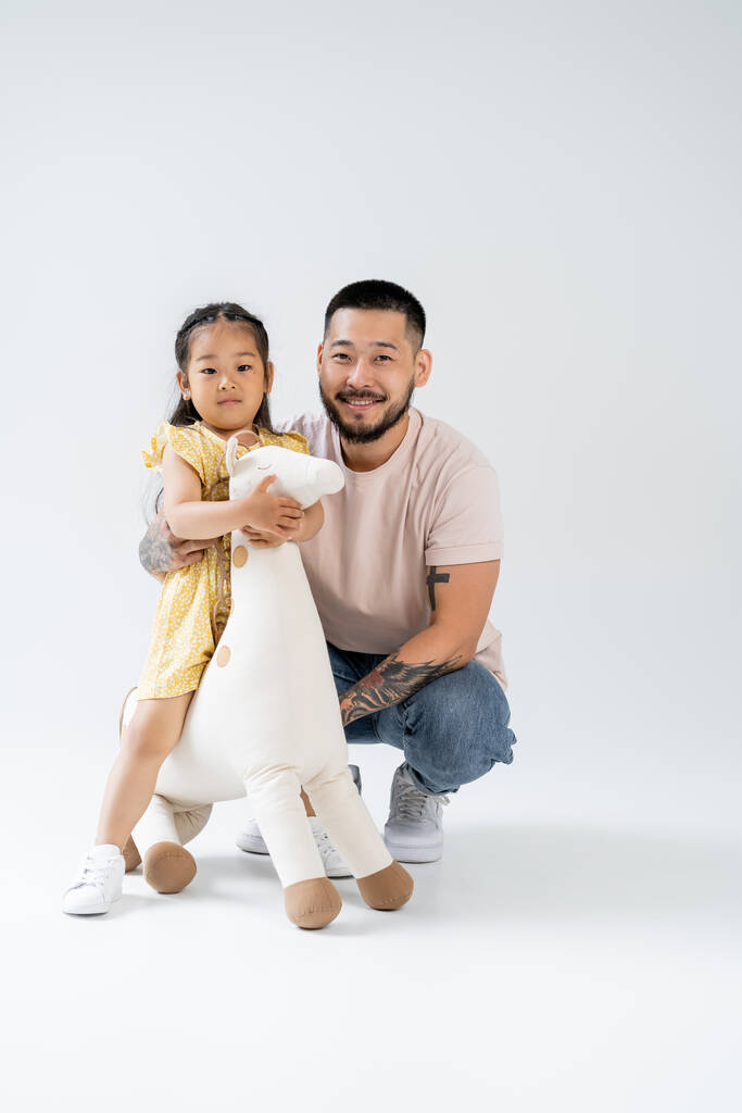 полная длина счастливого азиатского ребенка, держащего игрушечного жирафа и стоящего рядом с татуированным отцом на сером - Фото, изображение