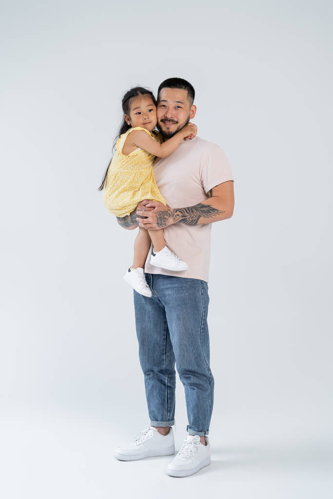 volle Länge der glücklichen und tätowierten asiatischen Vater hält lächelnde Vorschultochter auf grau - Foto, Bild