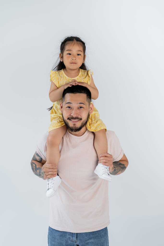feliz asiático niño sentado en hombros de tatuado padre aislado en gris - Foto, Imagen