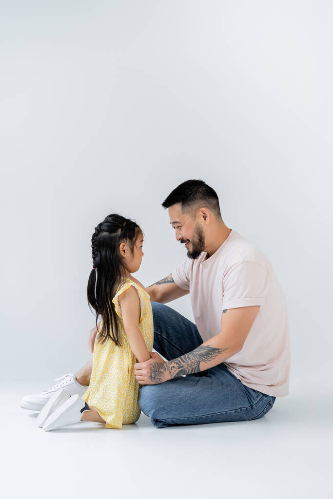 pełna długość szczęśliwy Azji dziecko siedzi w pobliżu wesoły ojciec w dżinsy na szary - Zdjęcie, obraz