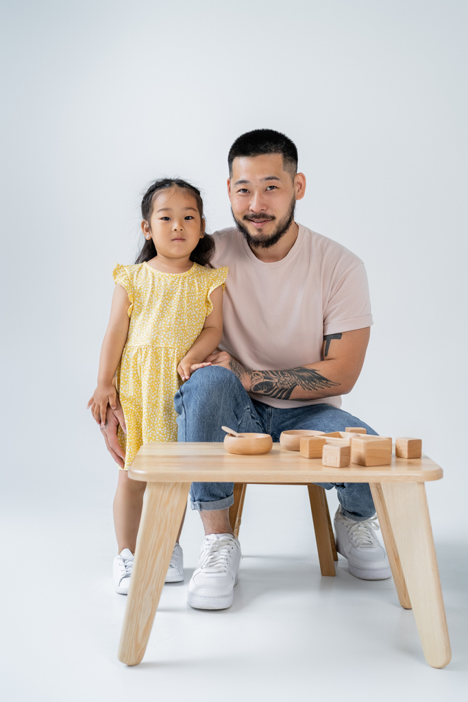 wesoły i wytatuowany ojciec i azjatycka przedszkolanka córka przy stole z drewnianymi zabawkami na szarości - Zdjęcie, obraz