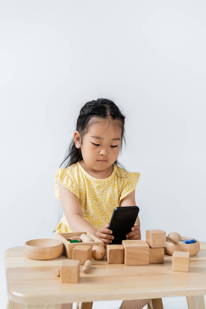 menina asiática usando smartphone perto de brinquedos de madeira na mesa isolado no cinza - Foto, Imagem