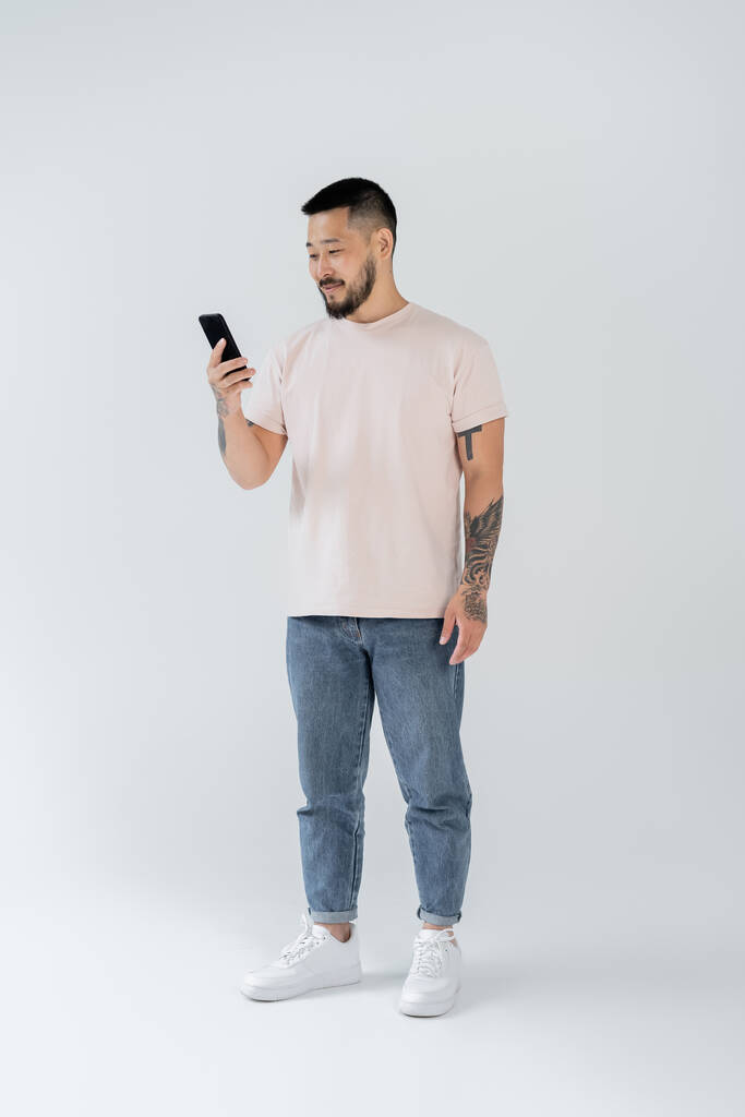 volledige lengte van lachende aziatische man met tatoeages met smartphone op grijs - Foto, afbeelding