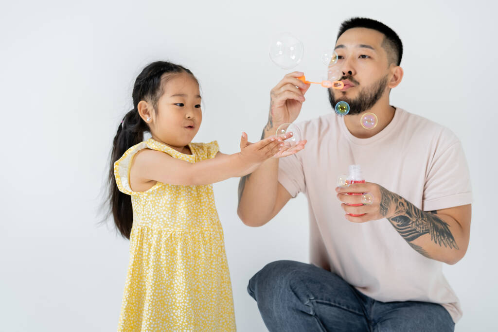 Asijské otec foukání mýdlo bubliny v blízkosti ohromený předškolák dcera izolované na šedé - Fotografie, Obrázek