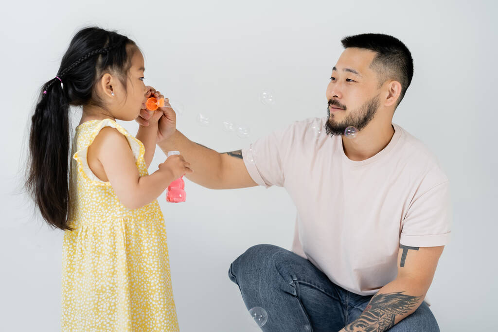 азіатський дошкільник дитина в жовтій сукні дме мильні бульбашки біля татуйованого батька на сірому
 - Фото, зображення