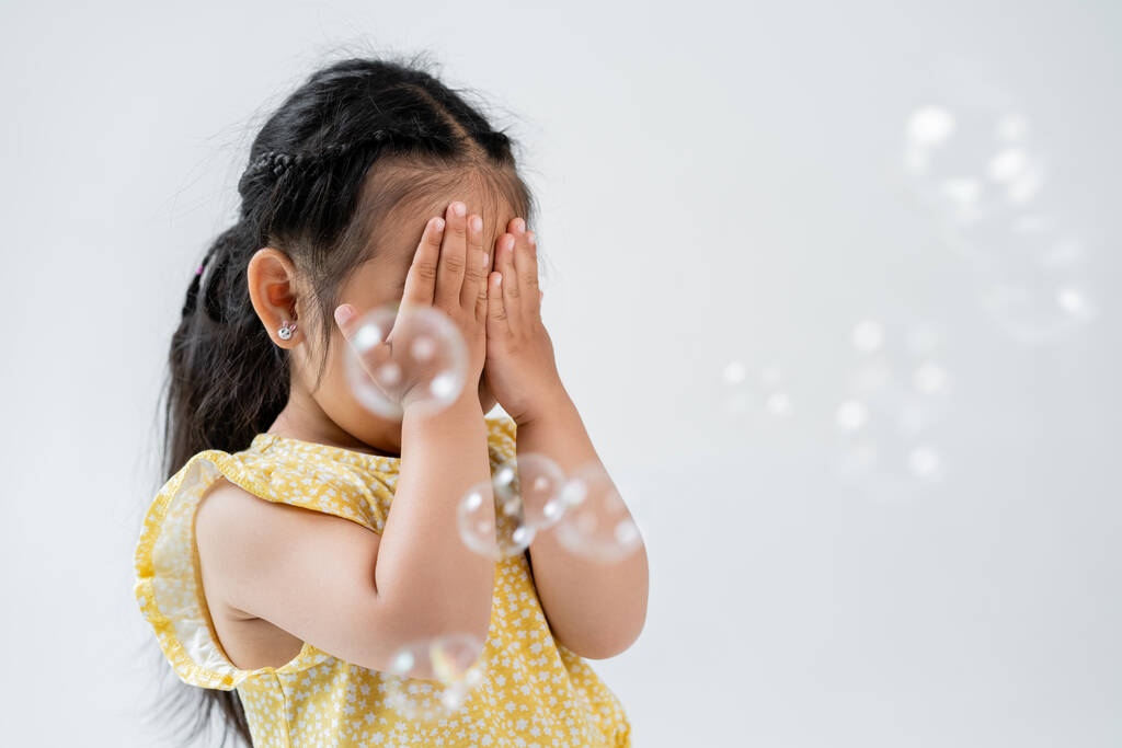retrato de niña preescolar asiática en vestido amarillo cubriendo la cara con las manos cerca de pompas de jabón aisladas en gris  - Foto, Imagen