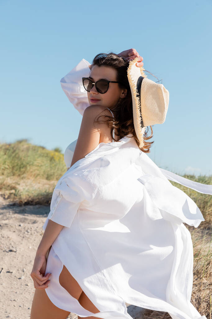 mujer joven en gafas de sol y camisa blanca sosteniendo sombrero de paja en la playa de arena - Foto, Imagen