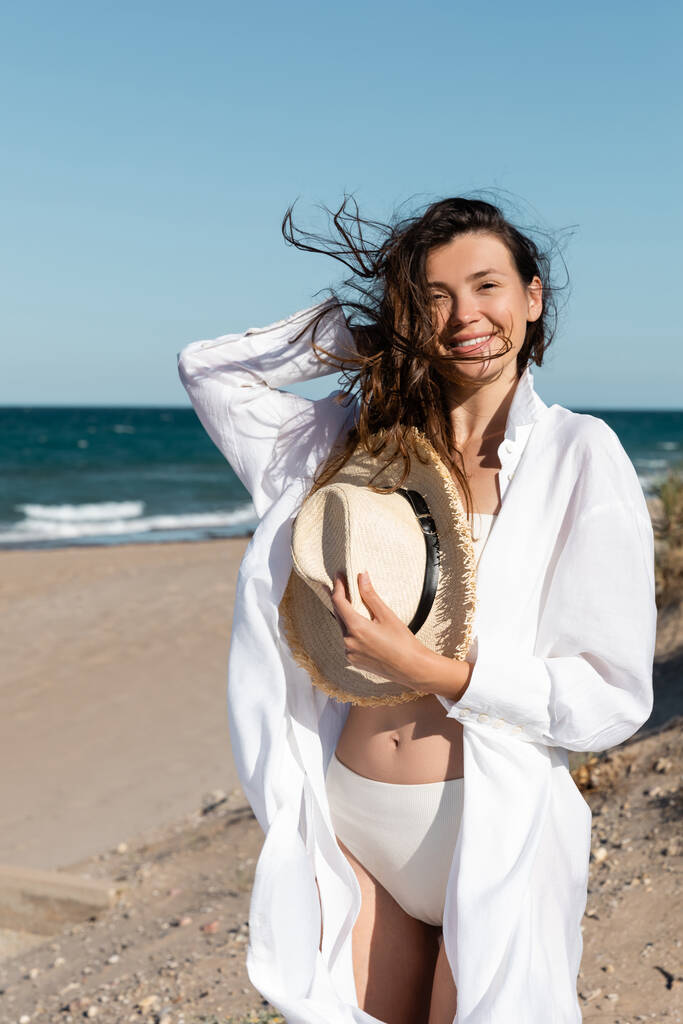 Beyaz gömlekli ve mayo giyen neşeli genç bir kadın sahile yakın bir yerde hasır şapka tutuyor. - Fotoğraf, Görsel