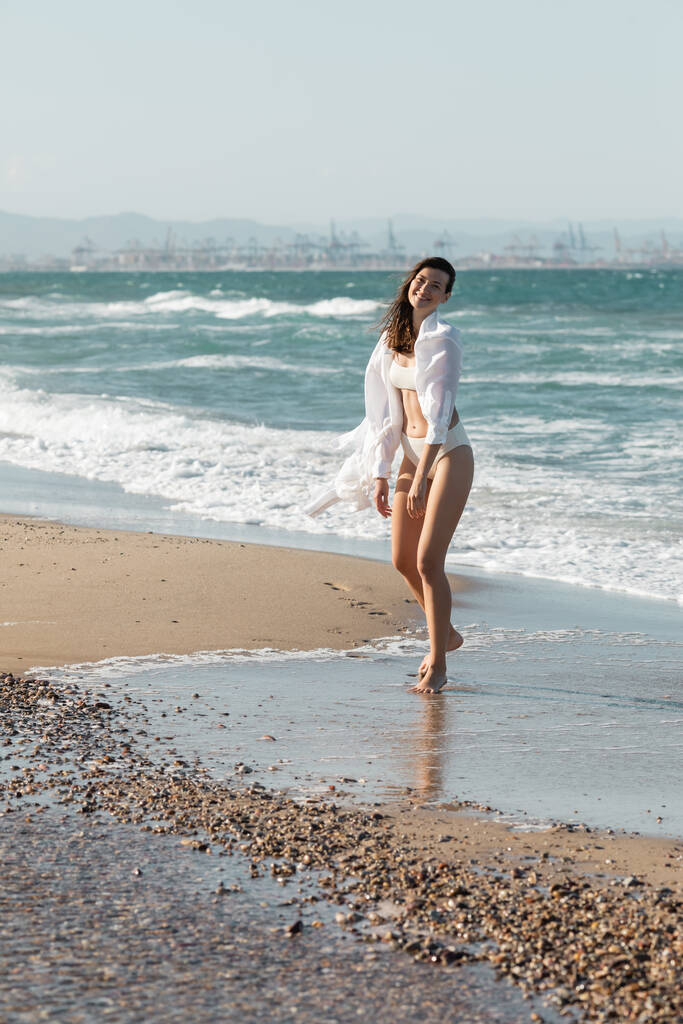 volle Länge einer glücklichen jungen Frau in weißem Hemd und Badebekleidung, die am Ufer des Meeres spaziert  - Foto, Bild