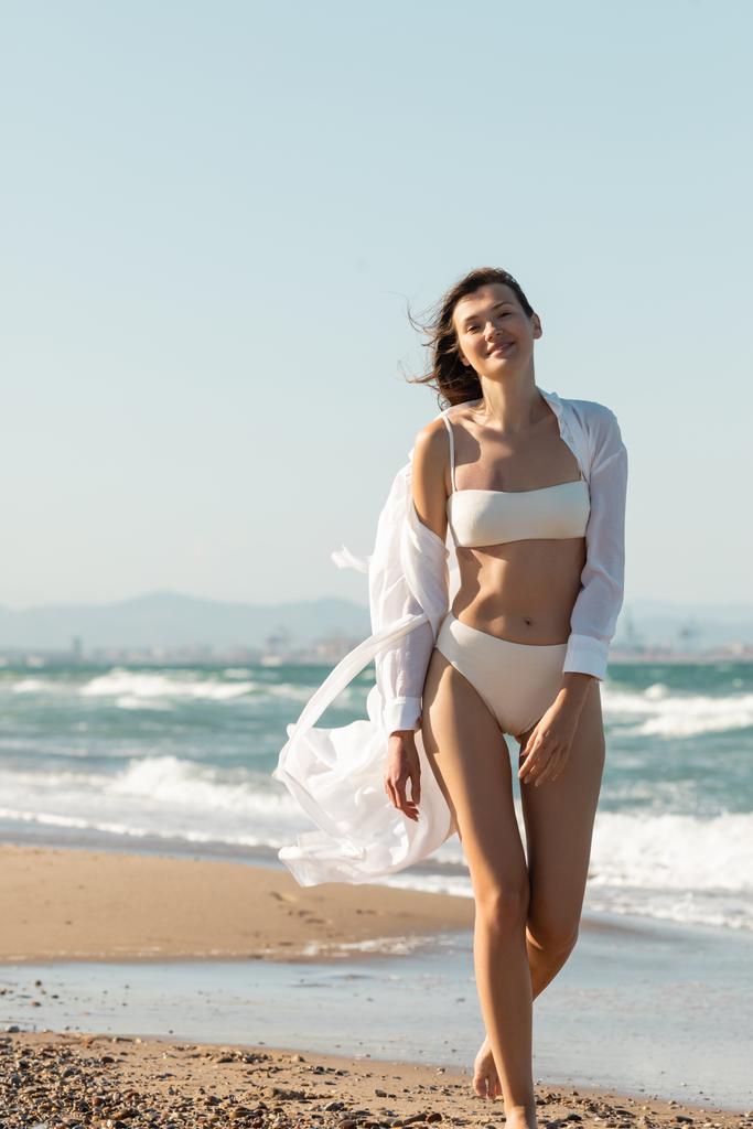 happy young woman in white shirt and swimwear walking on sea shore  - Photo, Image