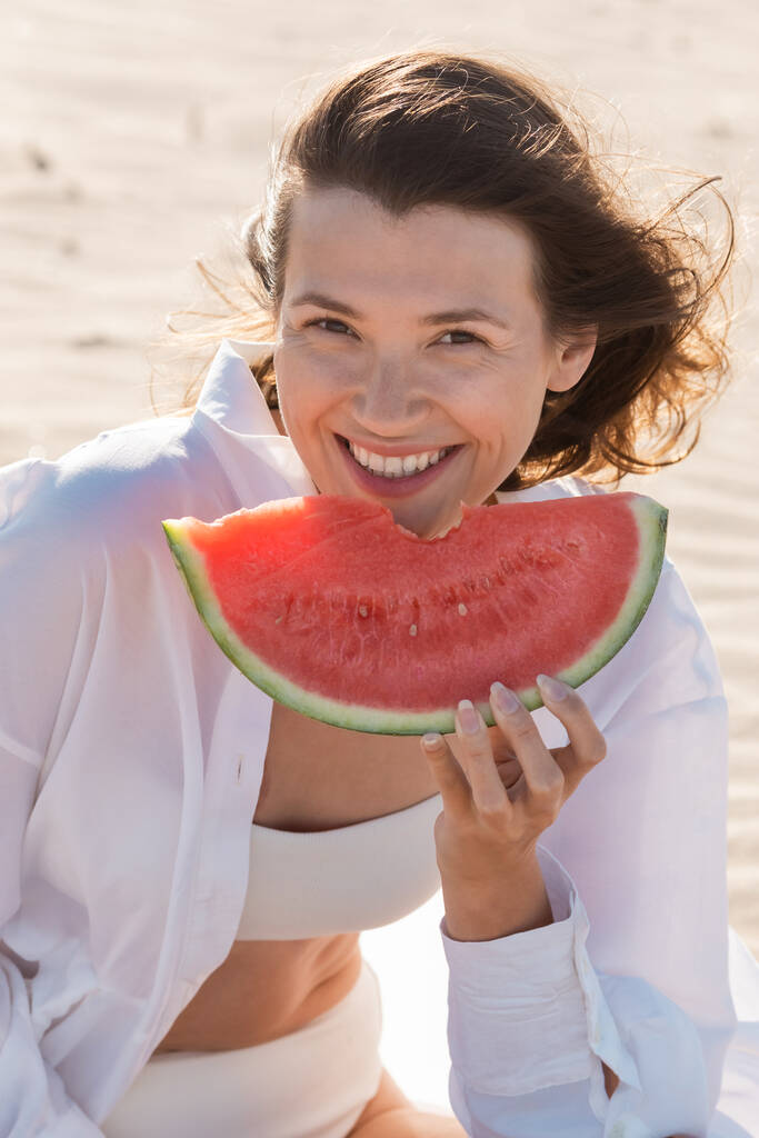 Porträt einer fröhlichen jungen Frau im weißen Hemd mit leckerer Wassermelone am Sandstrand - Foto, Bild