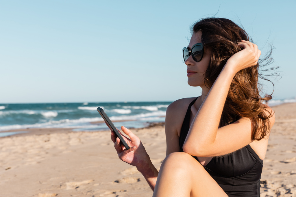 młoda kobieta w stroju kąpielowym i okularach przeciwsłonecznych trzyma smartfona w pobliżu morza - Zdjęcie, obraz