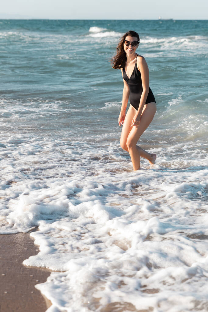 黒い水着の陽気な女性と海に立つスタイリッシュなサングラス - 写真・画像